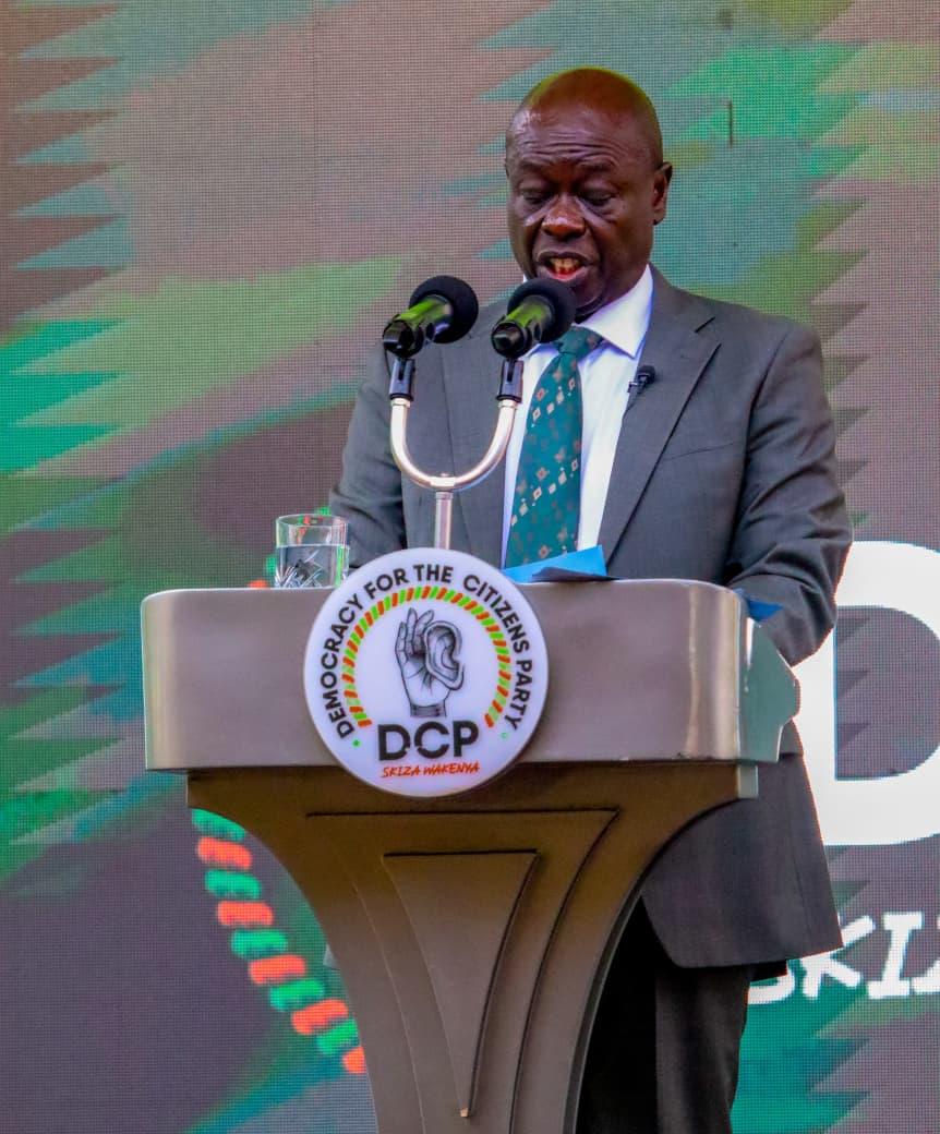 Rigathi Gachagua in a grey suit, speaking at a podium with the DCP seal